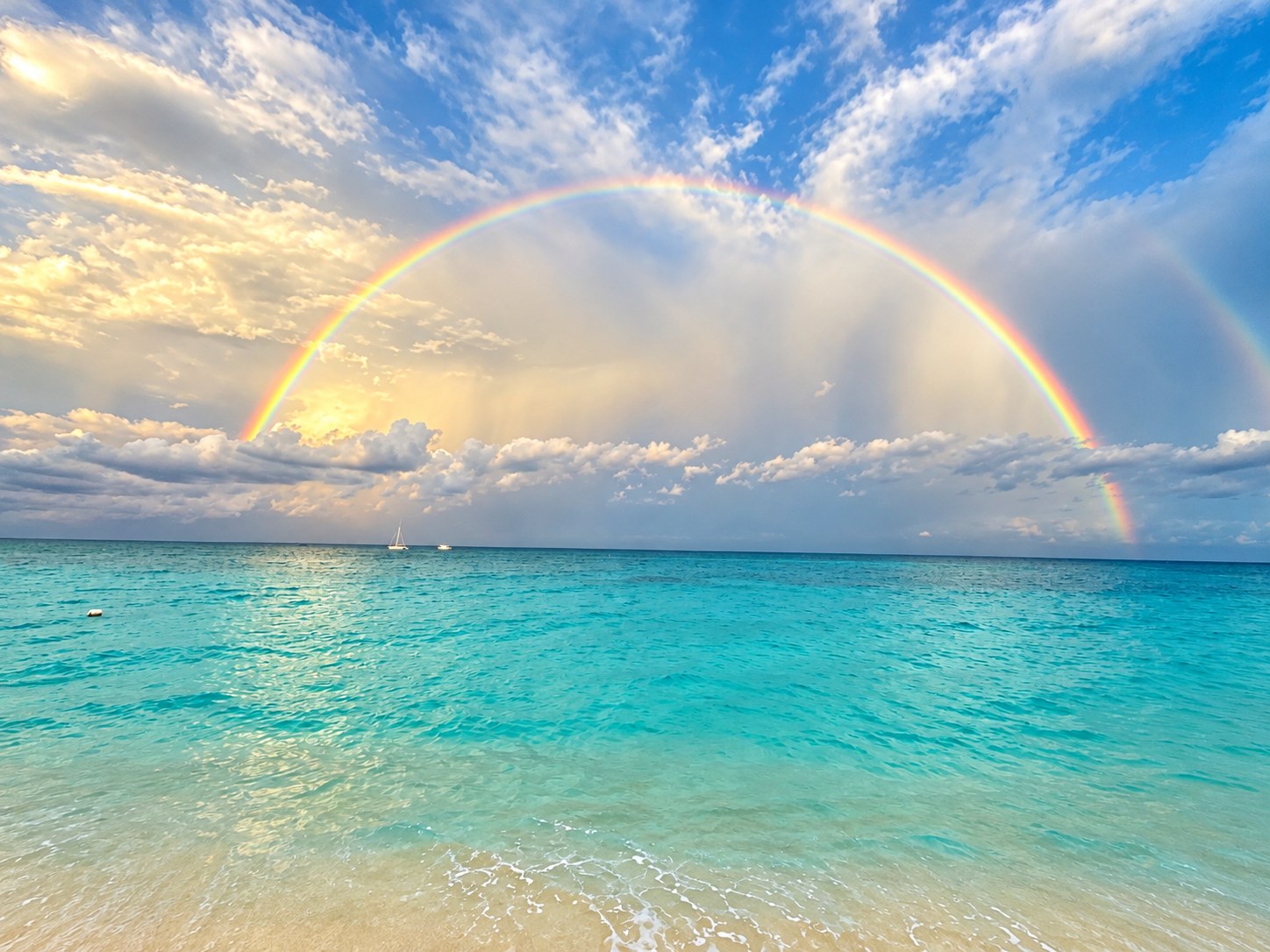 A full rainbow arcing over a turquoise tropical beach with a sailboat on the horizon